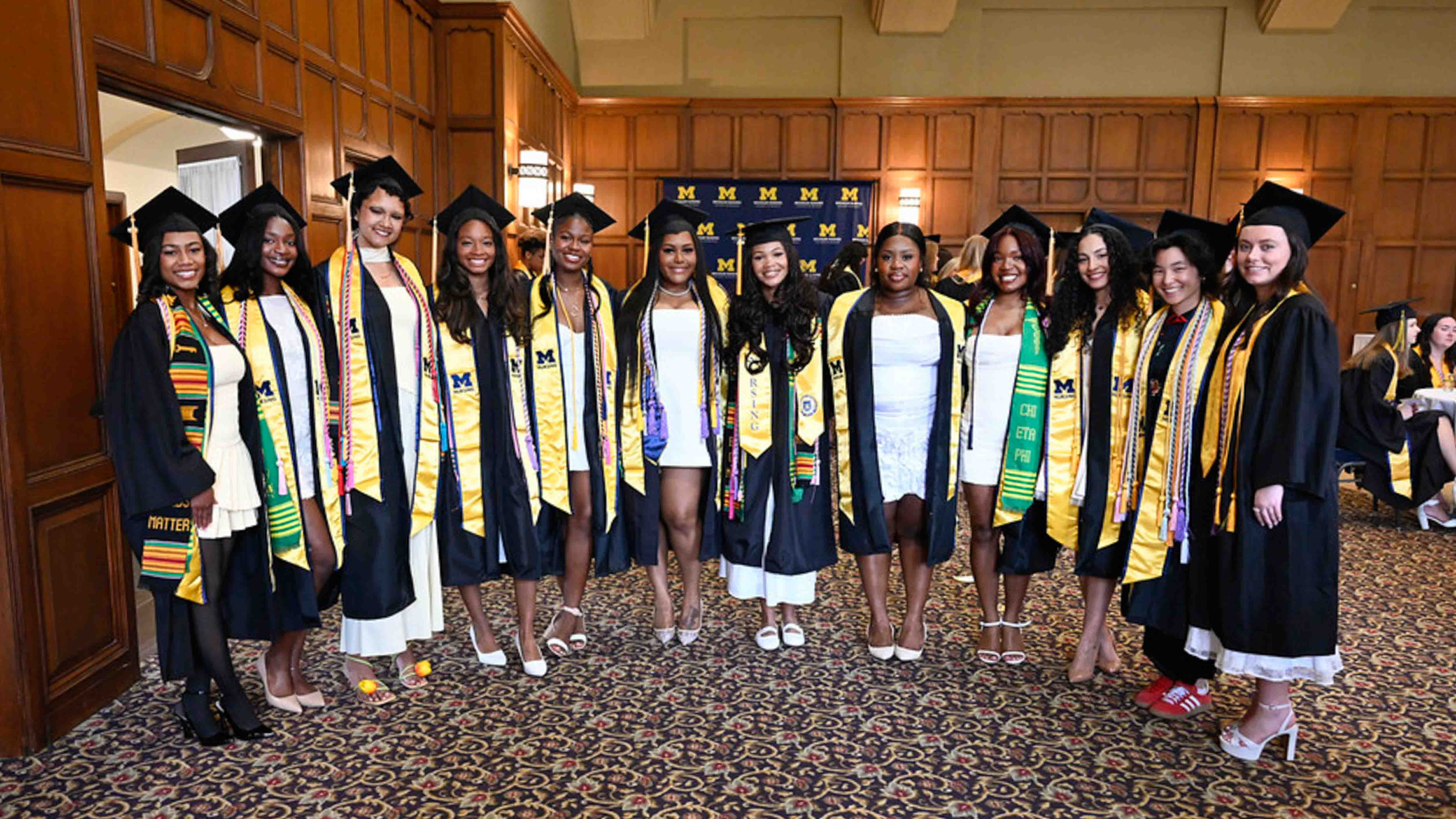 A group of nursing graduates in caps, gowns, and tassels posing in a line together at Hill Auditorium, smiling at the camera.