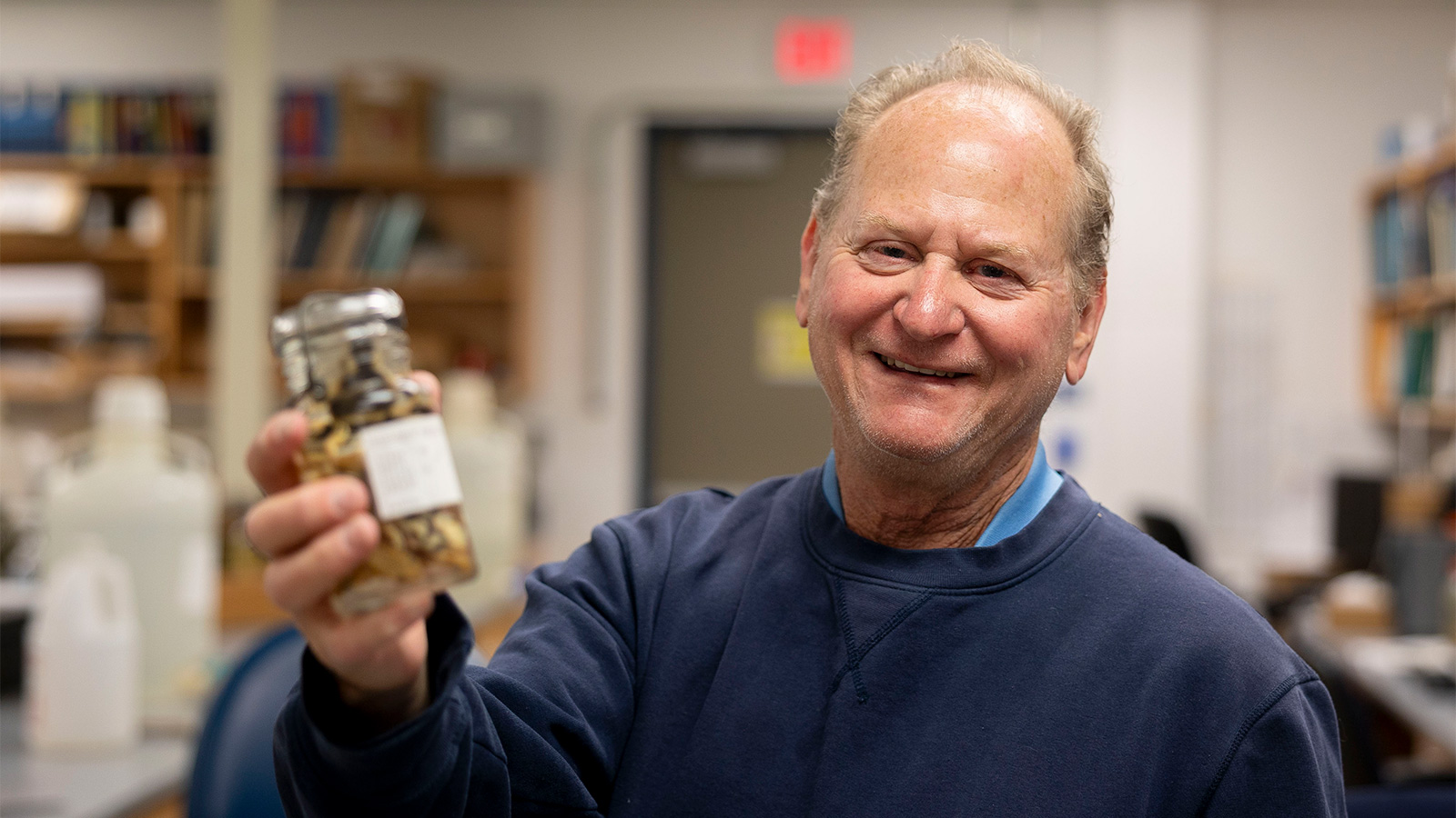 Greg Schneider wearing a navy blue sweatshirt and blue collared shirt holds a glass jar containing preserved specimens, standing in a laboratory lined with shelves and bottles.