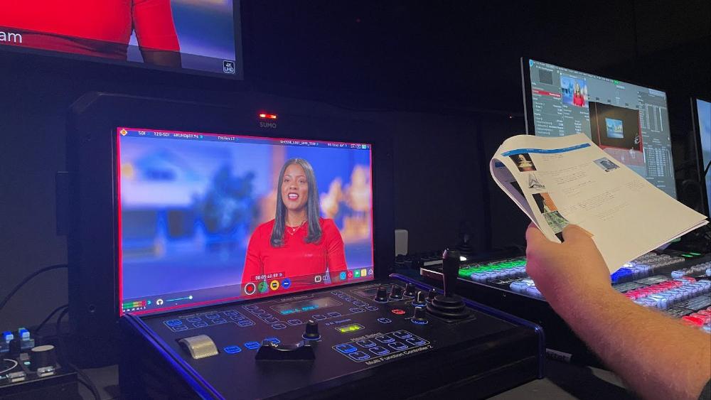 A woman appears on a screen attached to a control panel that is surrounded by other digital video production equipment. The hand of someone who is cropped out of the photo is holding a packet of papers.