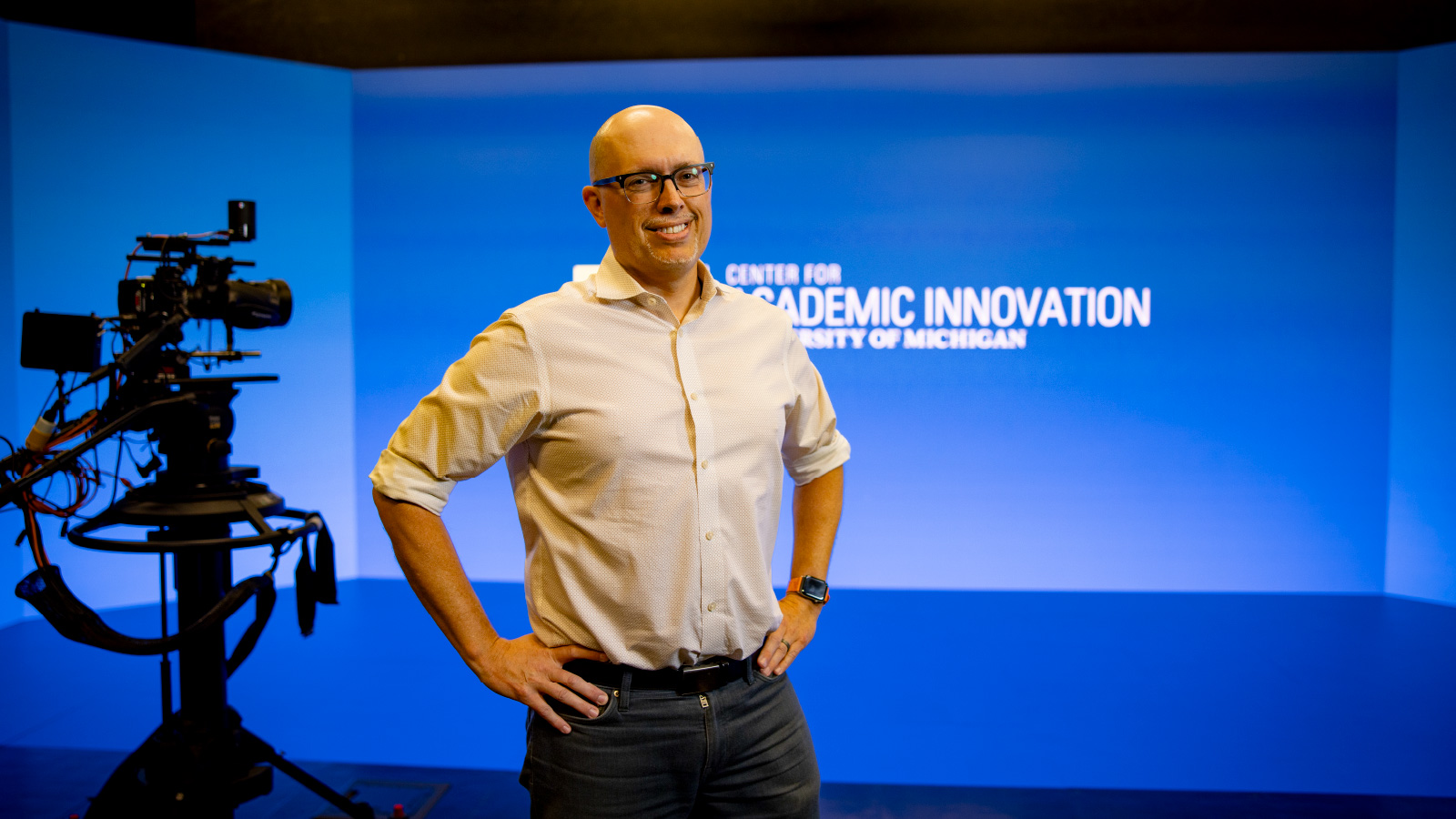 A man stands with his hands on his hips in front of a blue screen that reads “Center for Academic Innovation.” A movie camera is to his right.