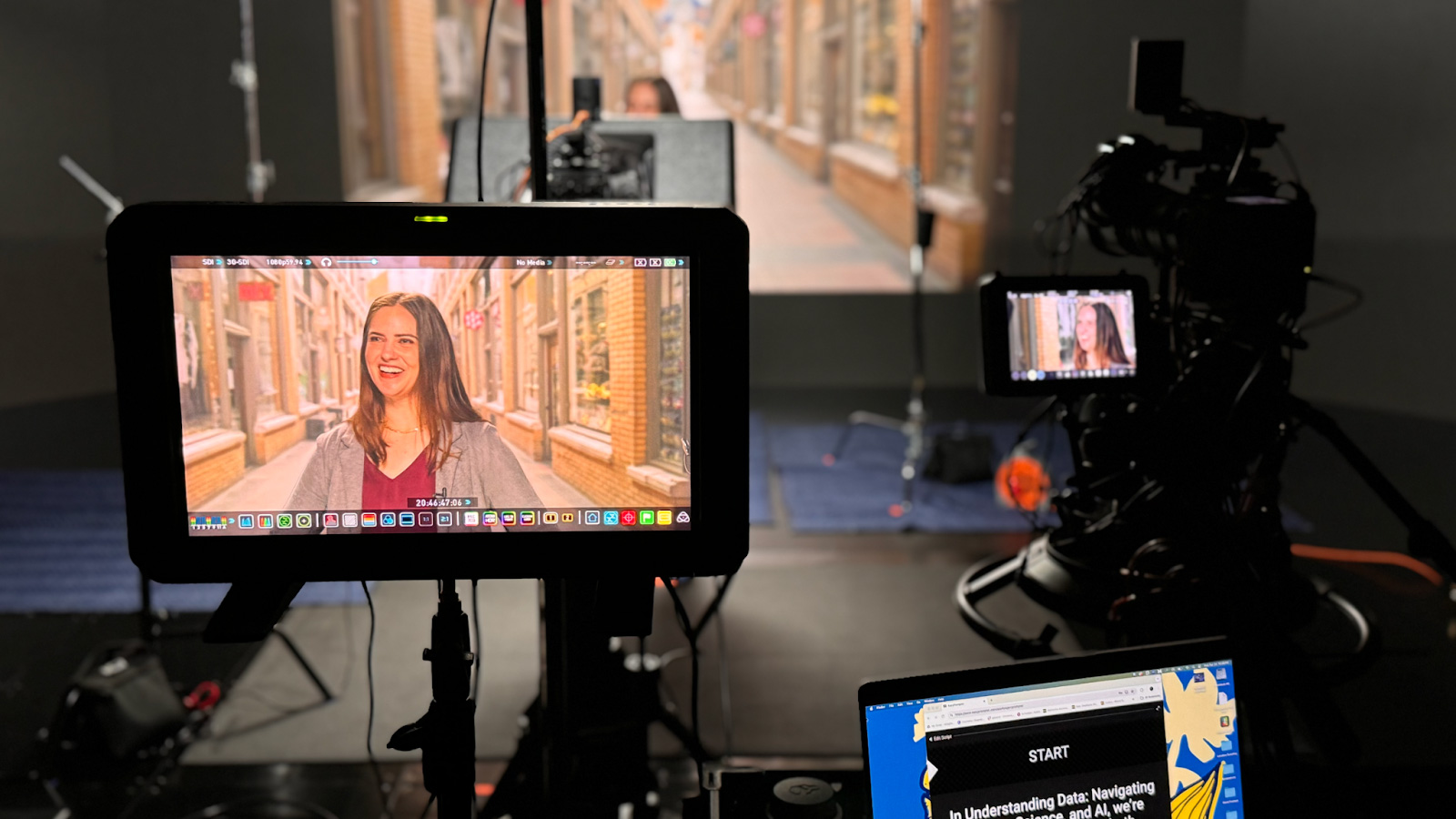 A woman is standing on set in front of several cameras for filming. Her likeness is visible in two different monitors attached to film equipment in the foreground of the photo.