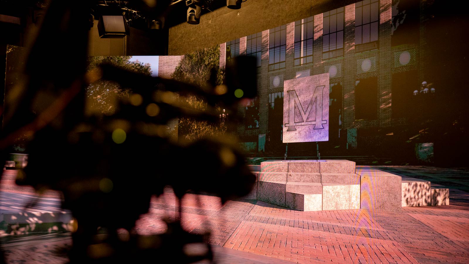 A large screen is displaying a video image of the Diag in front of Hatcher Library. A Block M engraved in stone hovers above the bricks. A film camera appears out of focus in the foreground. 