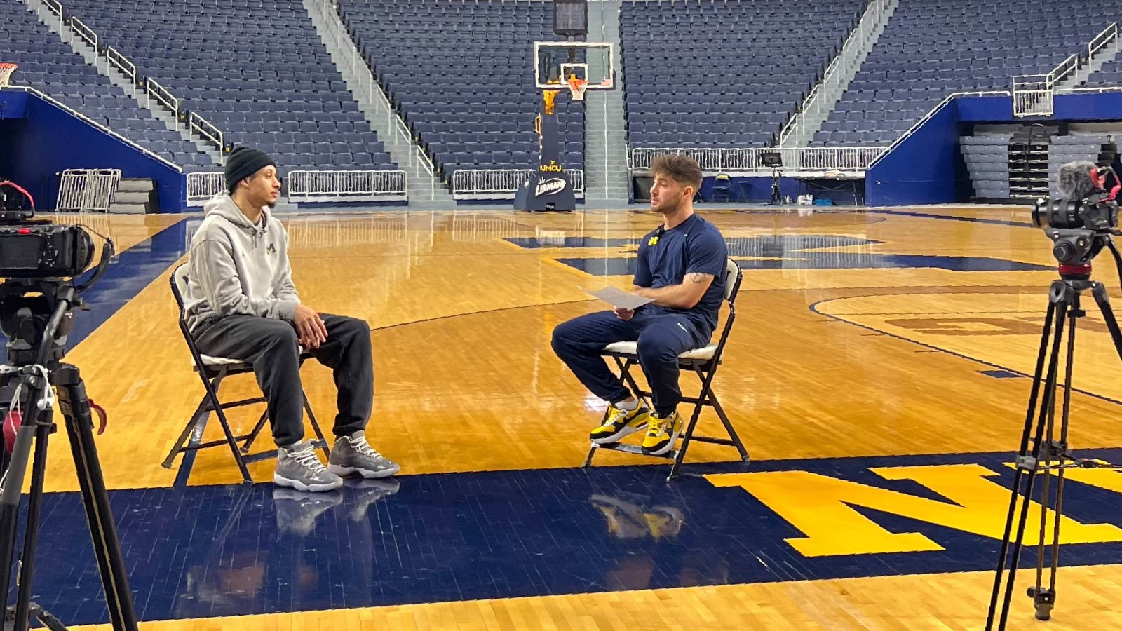 Uriel Zeitz and Jaelin Llewellyn sitting in chairs, face to face, in the middle of an empty basketball court. 