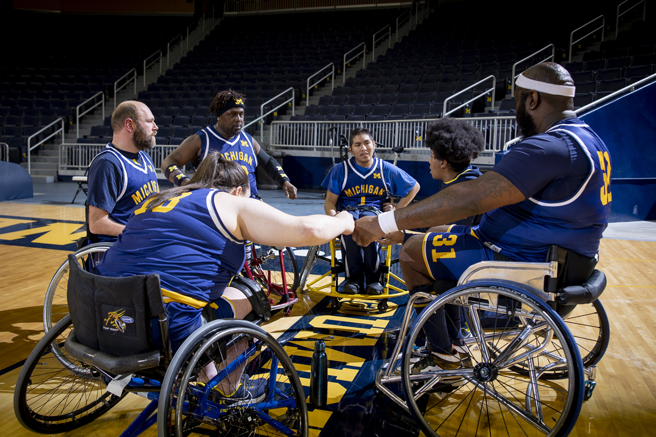 Men and women in wheelchairs and basketball uniforms huddled together in a circle, fist bumping.