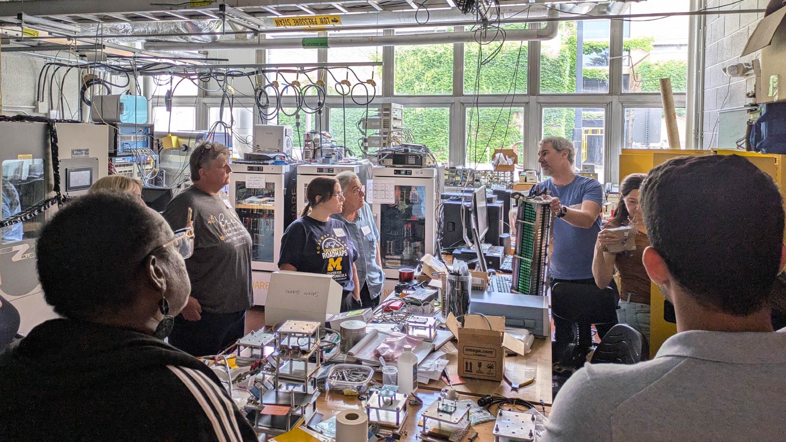 A group of people watch as someone demonstrates a piece of machinery. In the middle of the group, there is a large table covered in different components.