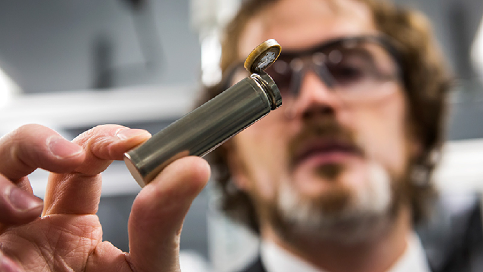 A person wearing safety glasses is holding a casing for a tube-shaped battery.