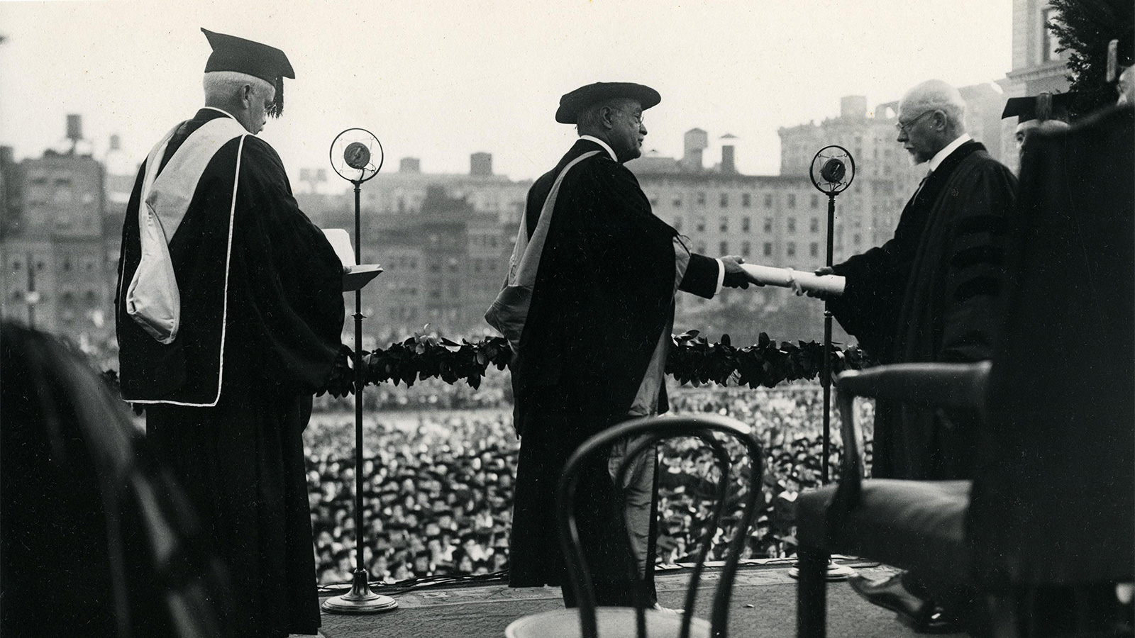A formal graduation ceremony with individuals in academic robes, one handing a diploma to another on a stage with a large crowd in the background.