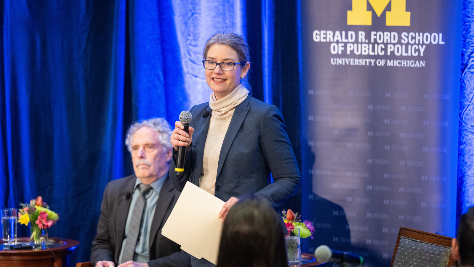  A woman standing holding a microphone with a man in the background and backdrop that reads “Gerald R. Ford School of Public Policy.”