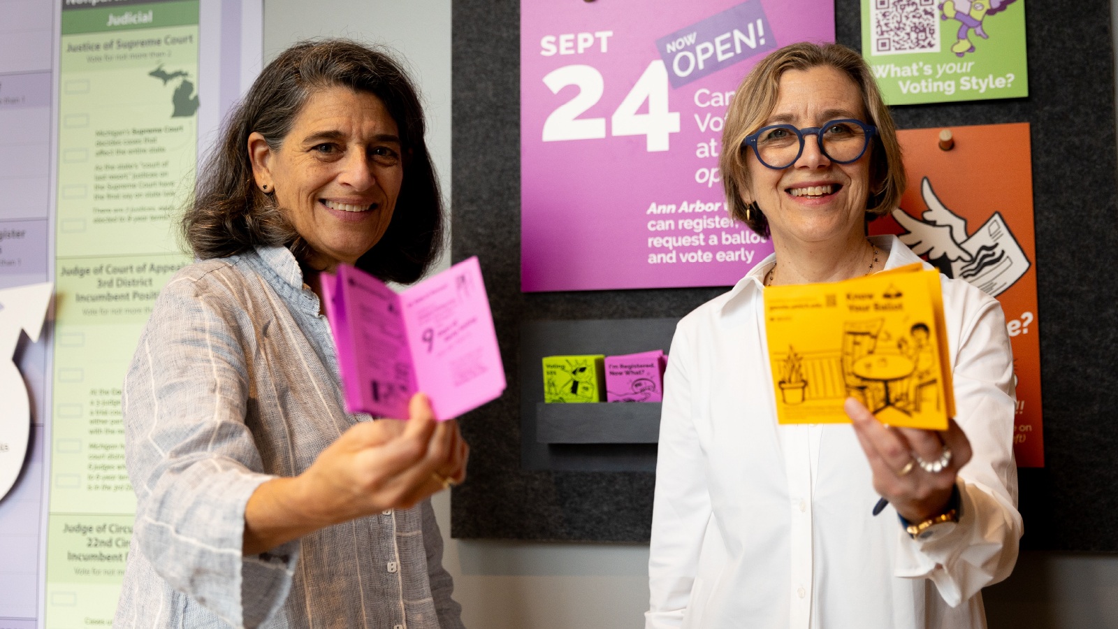 Two women standing in front of signs and posters encouraging voting in the 2024 election, holding up informational pamphlets. 