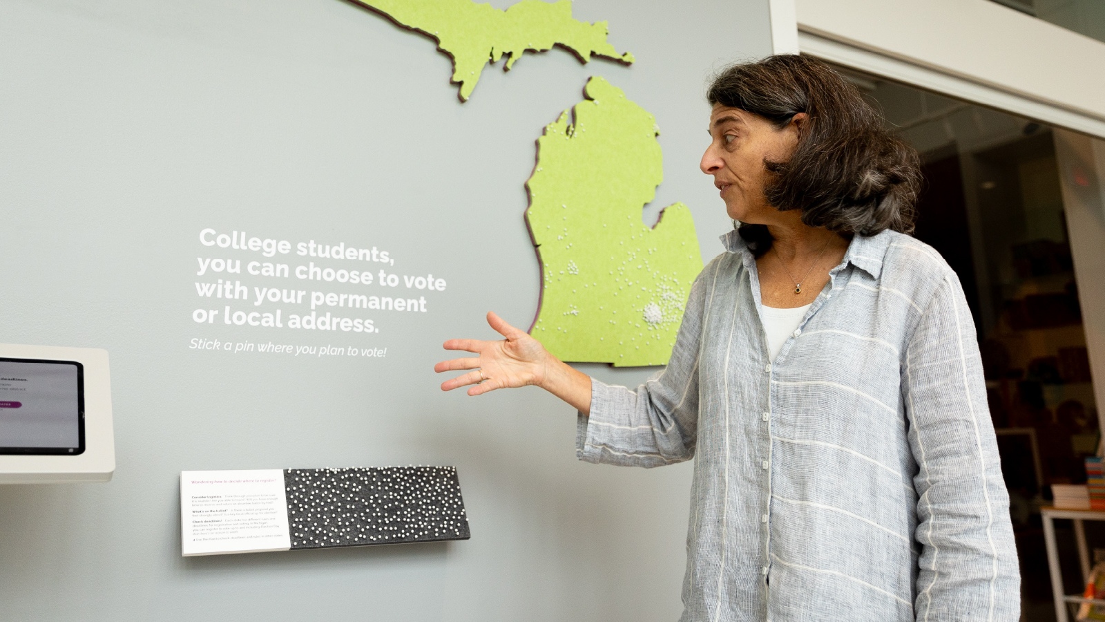 A woman standing in front of an artistic display of the state of Michigan, gesturing at instructions about voting written on the wall. 