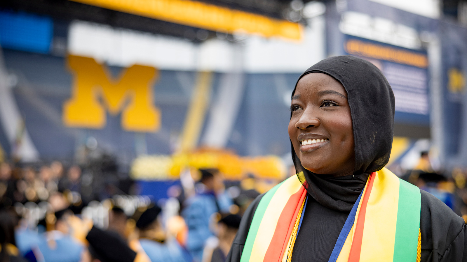 A person wearing academic regalia and a colorful stole stands in a graduation ceremony, with a blurred audience and a large block M in the background.