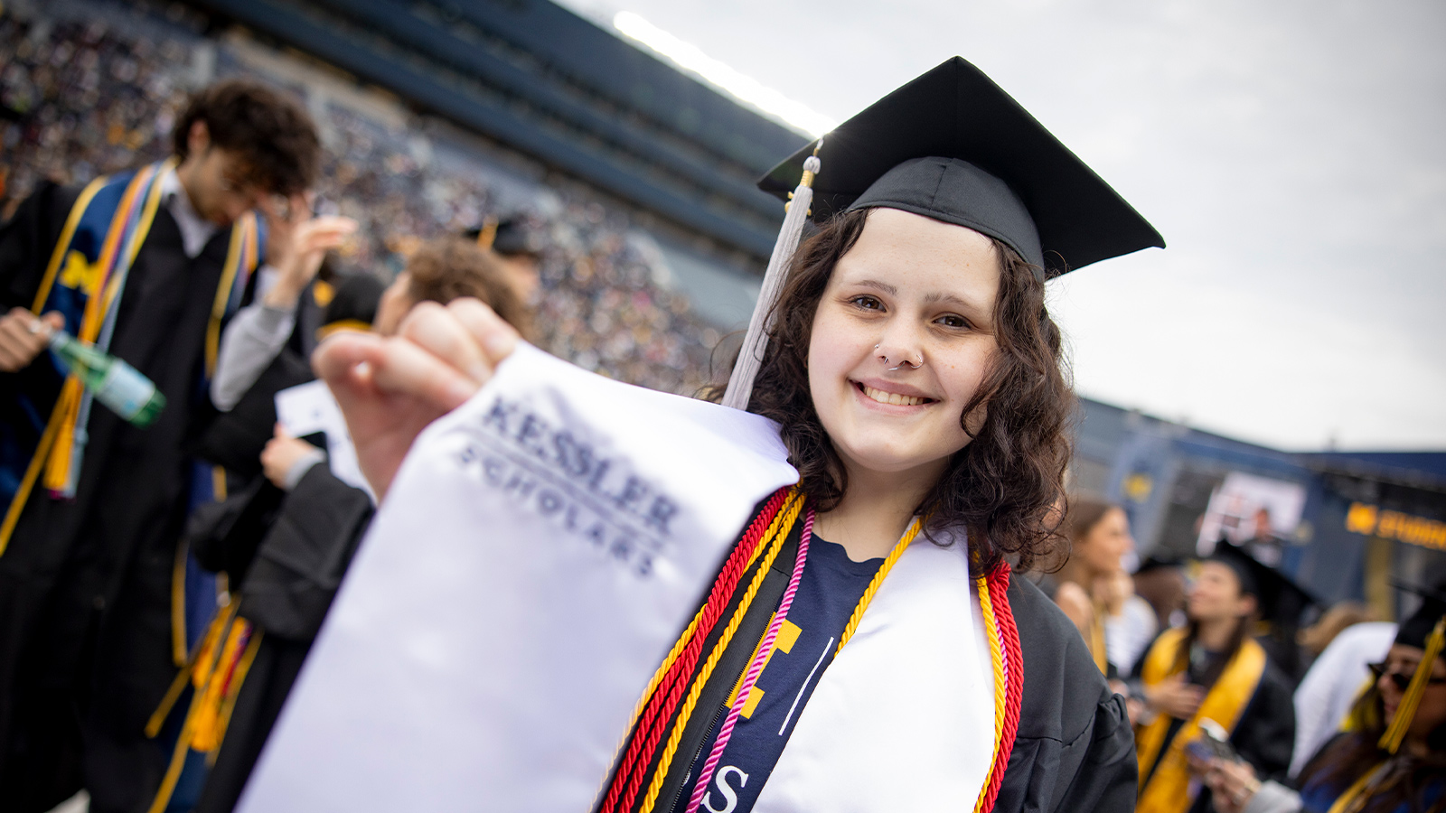 Graduate in cap and gown holds up a white stole with "Kessler Scholars" written on it, surrounded by others in academic regalia.
