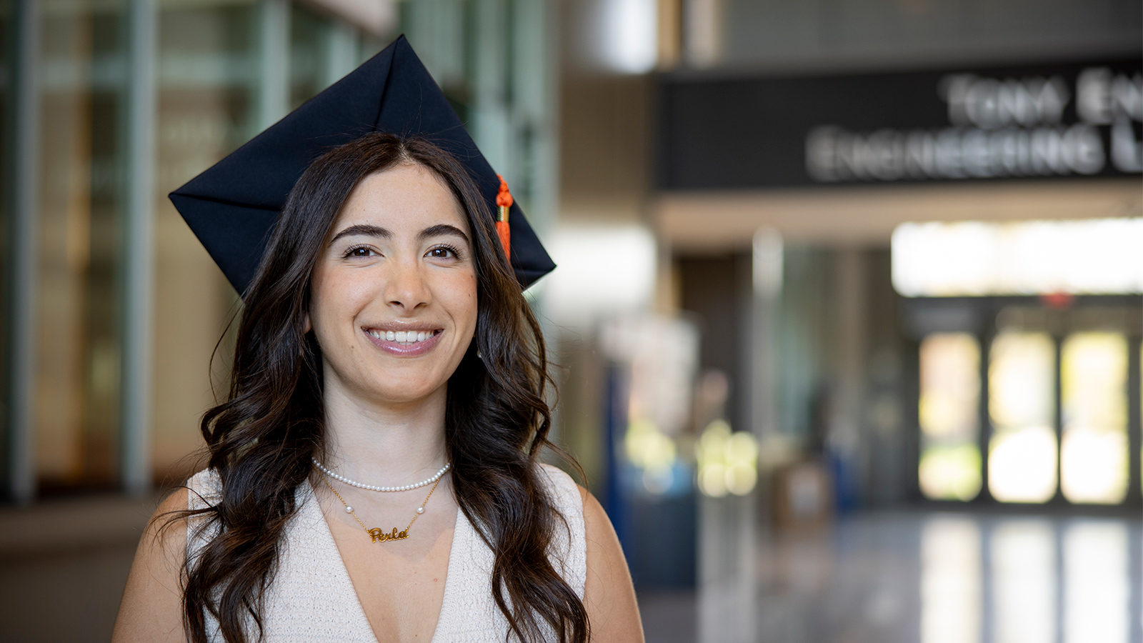 A person in a graduation cap and attire is seen indoors, wearing a pearl necklace, with the Tony England Engineering Lab sign in the background.