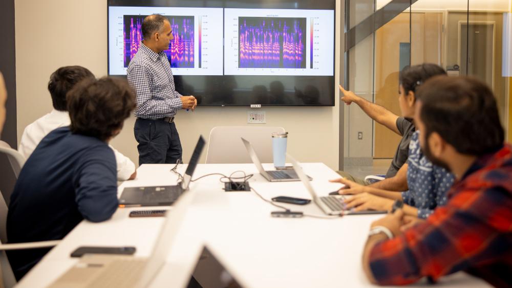 A man stands at the front of a room near a digital display screen. Several people are looking toward him. One of the people is pointing at the screen.