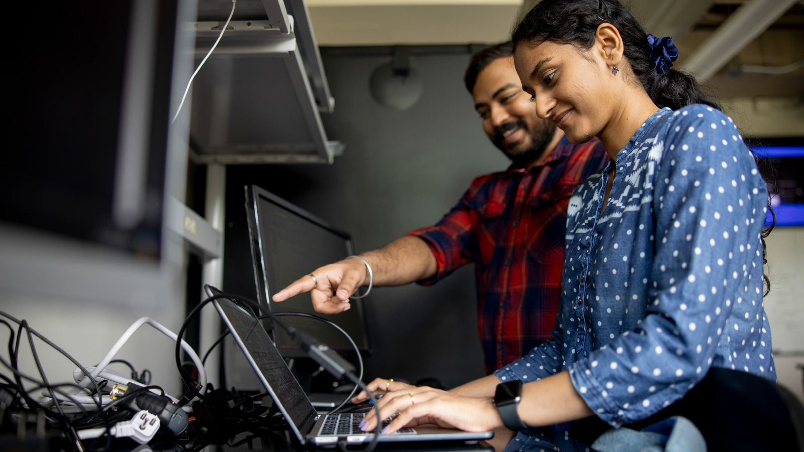 A man and a woman are standing next to each other at a laptop computer that is resting on a table covered in wires and other computer equipment.