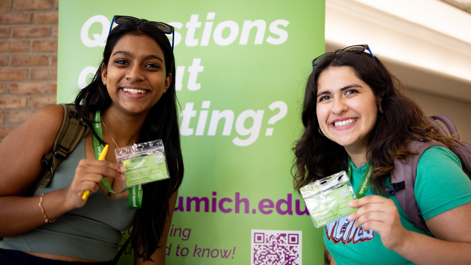 Two female students smiling and standing in front of a lime green sign that reads, “Questions about voting?”