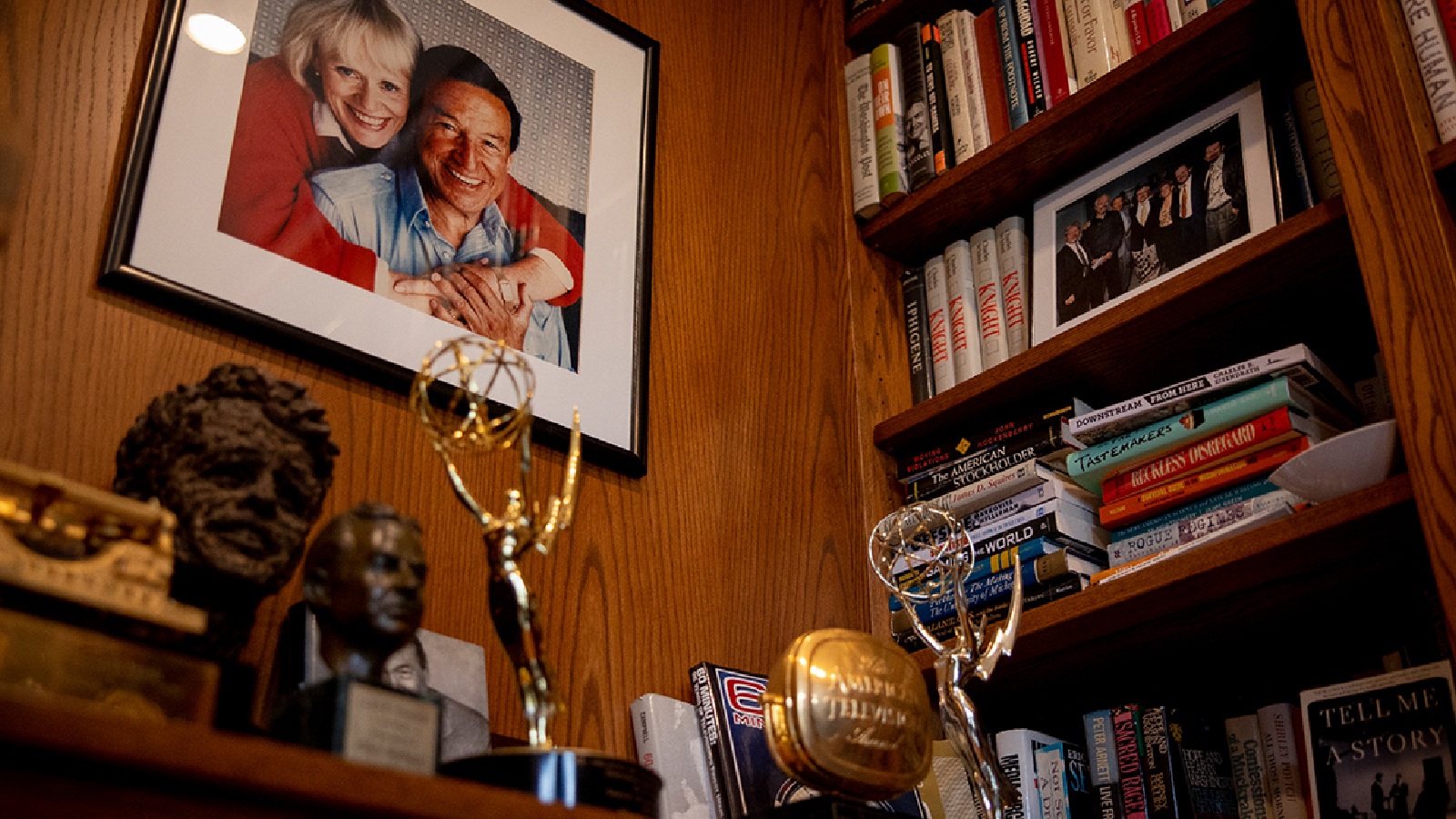 A close-up photo of a portrait of former journalist Mike Wallce and his wife hanging on a wall near a bookcase and a table filled with awards and statues.