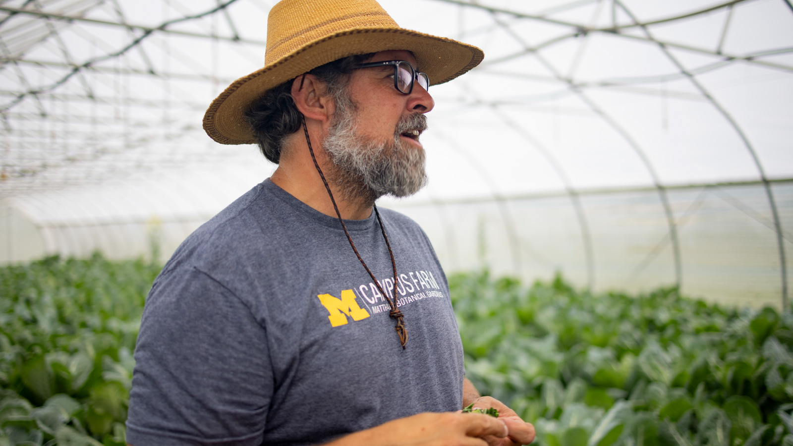  Campus Farm Program Manager Jeremy Moghtader standing in a greenhouse wearing a straw hat and blue T-shirt that says “M | Campus Farm.”