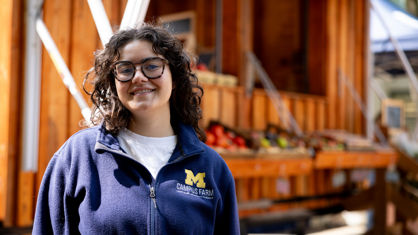 U‑M student Jenna Jarjoura stands in front of the U‑M Farm Stand. She is wearing a blue jacket that says “M Campus Farm.”