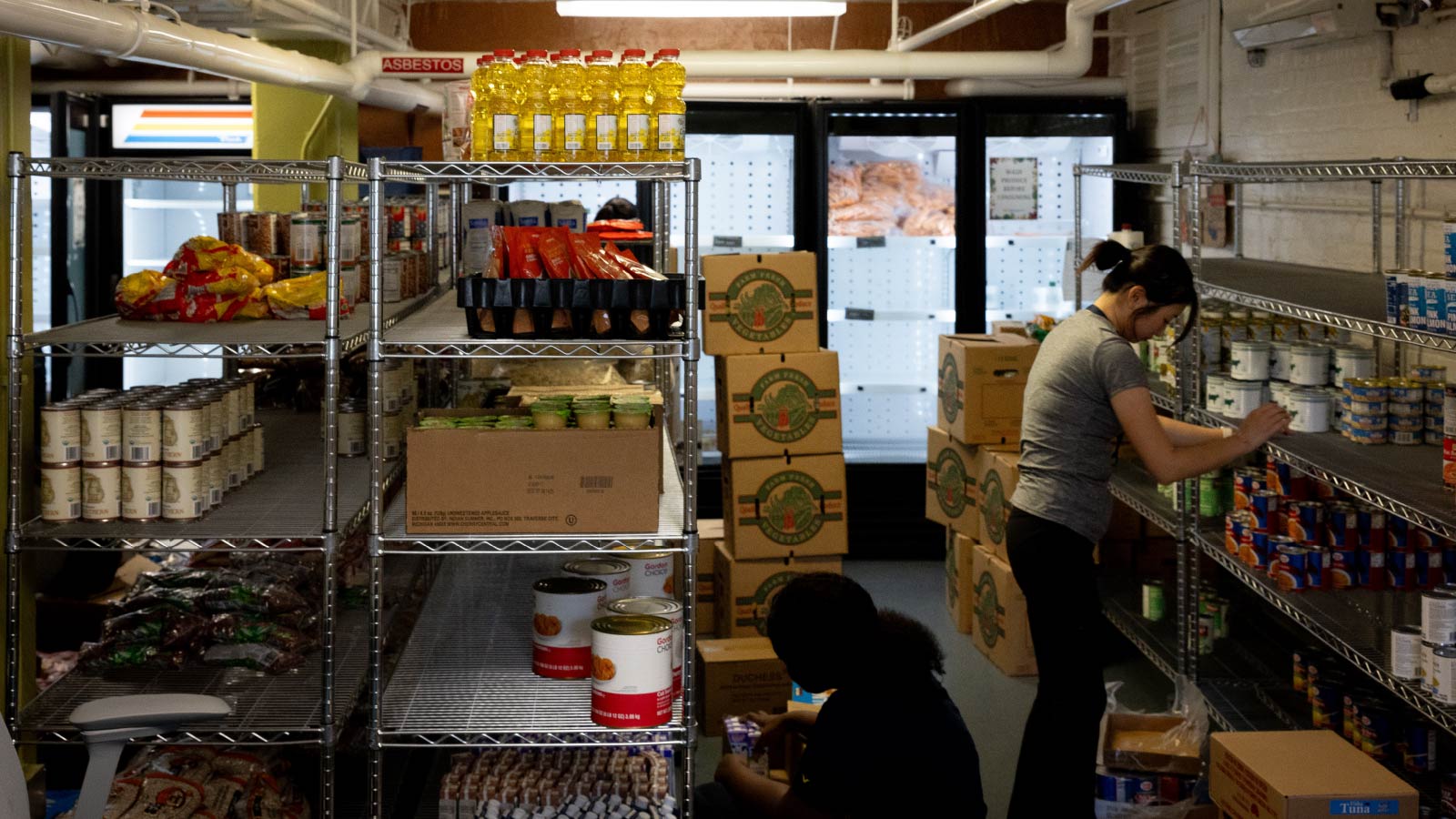 Two people stand between rows of shelves with food products stacked on them. There are boxes behind them and a row of coolers in the background.