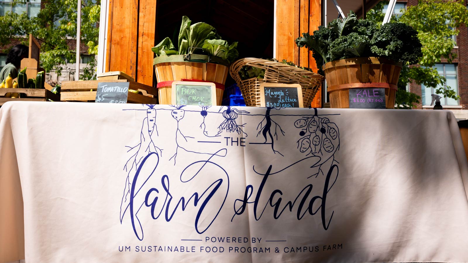 A table is covered with a tablecloth that says “The Farm Stand Powered by U‑M Sustainable Food Program & Campus Farm.” There are baskets of leafy green vegetables on the table. Part of a trailer is visible behind it.