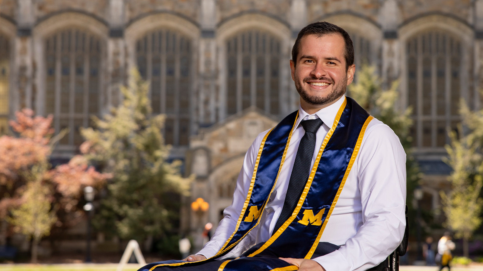 A person in a white shirt, black tie, and a blue stolen with maize trim and an "M" emblem sits in a wheelchair before a Gothic-style university building.