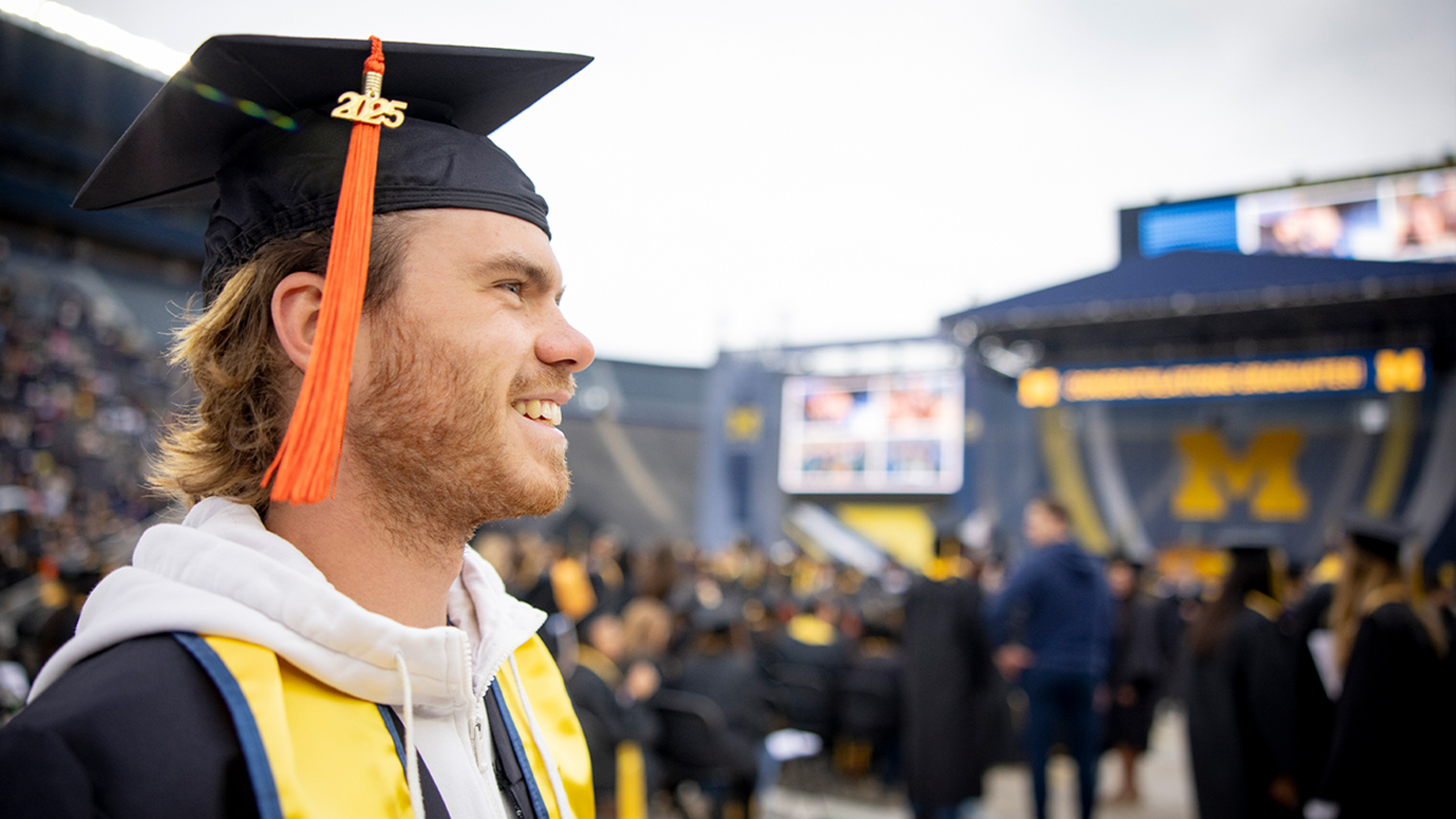 A graduate with a 2025 tassel and yellow stole stands in a stadium setting during a university commencement, with a large block M on stage in the background.