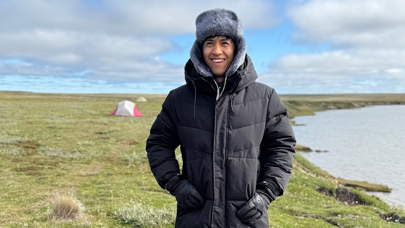  A man standing outside, in front of a large area of land and a body of water. He’s wearing a black coat and a gray fur trapper hat. 