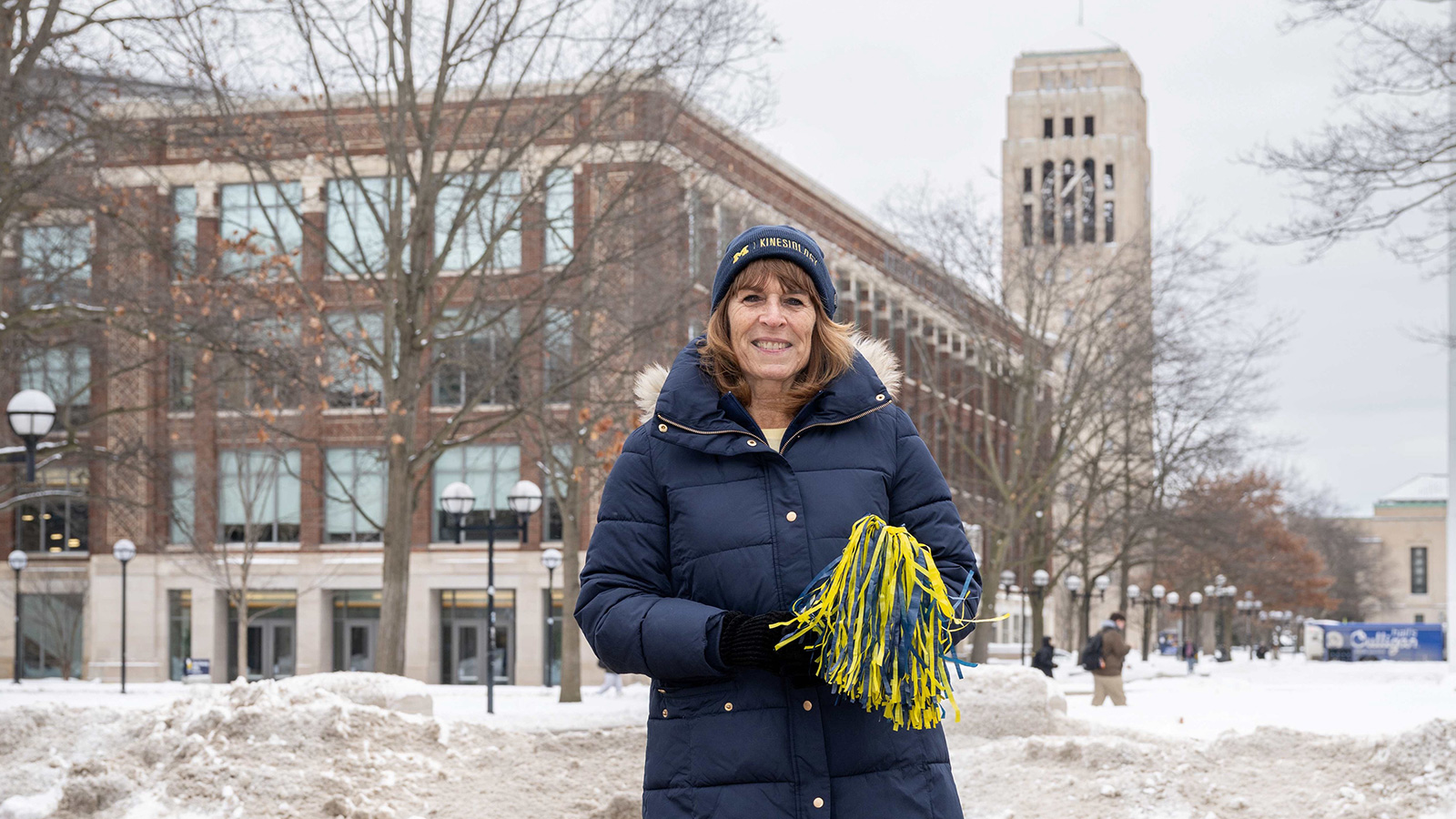 Pat in a winter coat standing outside in front of the University of Michigan School of Kinesiology, holding a yellow and blue pom-pom on a winter day.