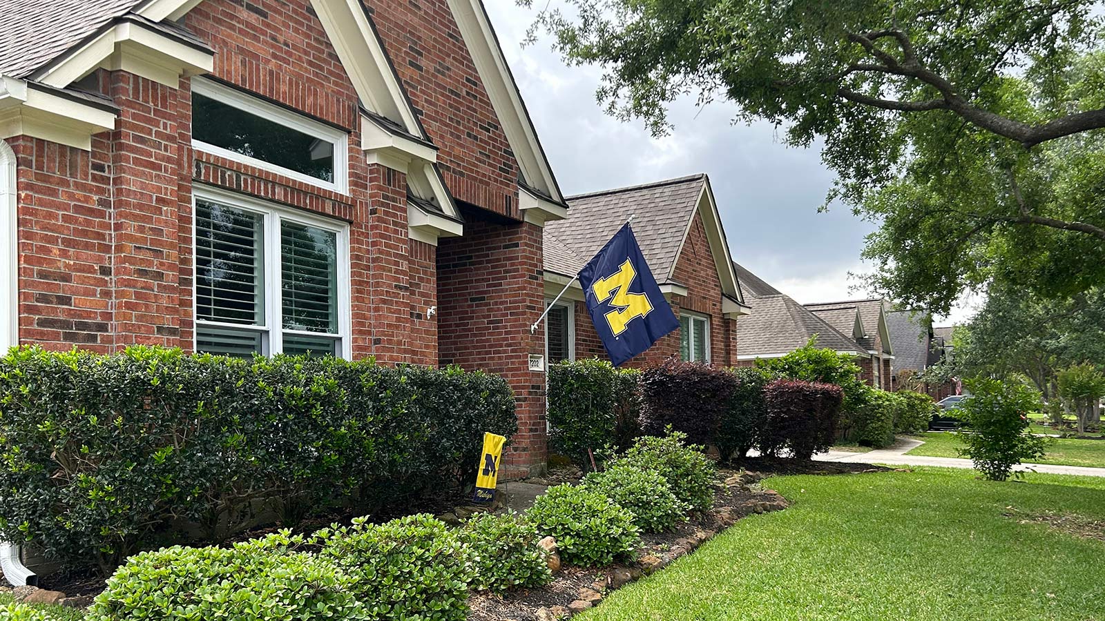 A brick house displays a University of Michigan flag and garden banner among green landscaping. 
