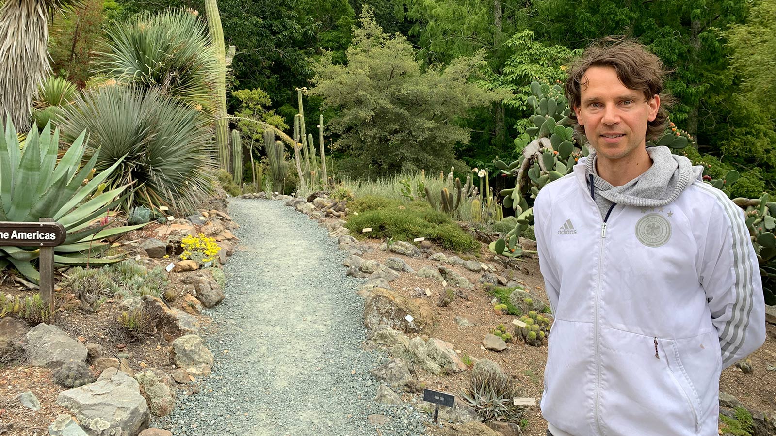 Roland Kersten standing on a path in a botanical garden, with trees, cacti, and other plants in the background.