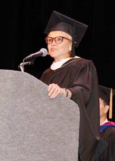 Joyce stands at a lectern. She is wearing a graduation cap and gown.