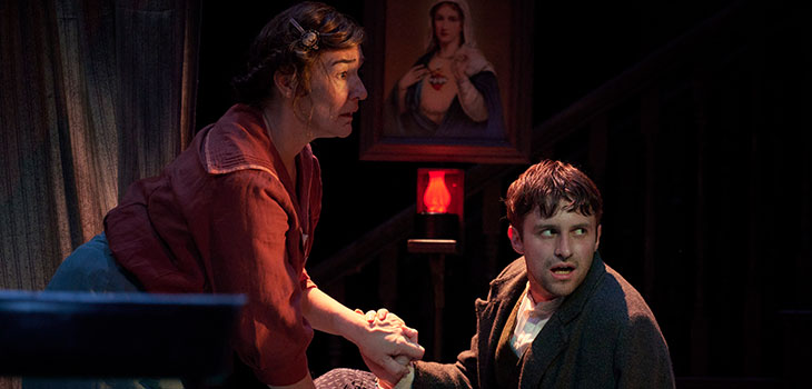 A woman gripping the hand of a seated man with concerned facial expressions.