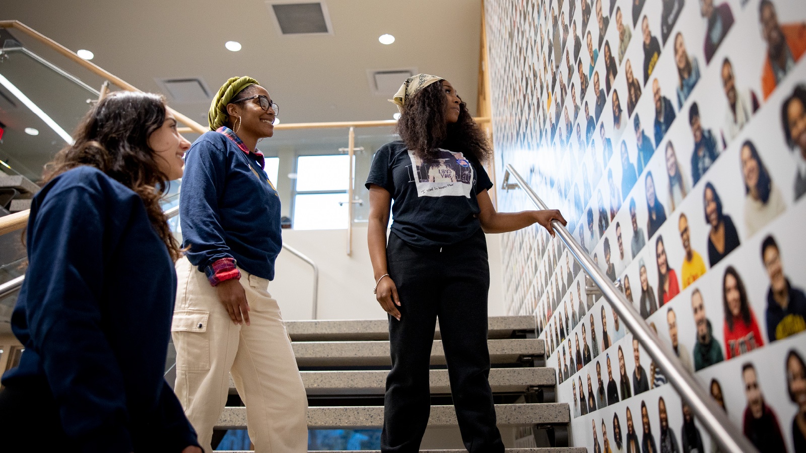 Three female students standing on a staircase at the Trotter Center looking at a wall with photos of different U‑M students. 