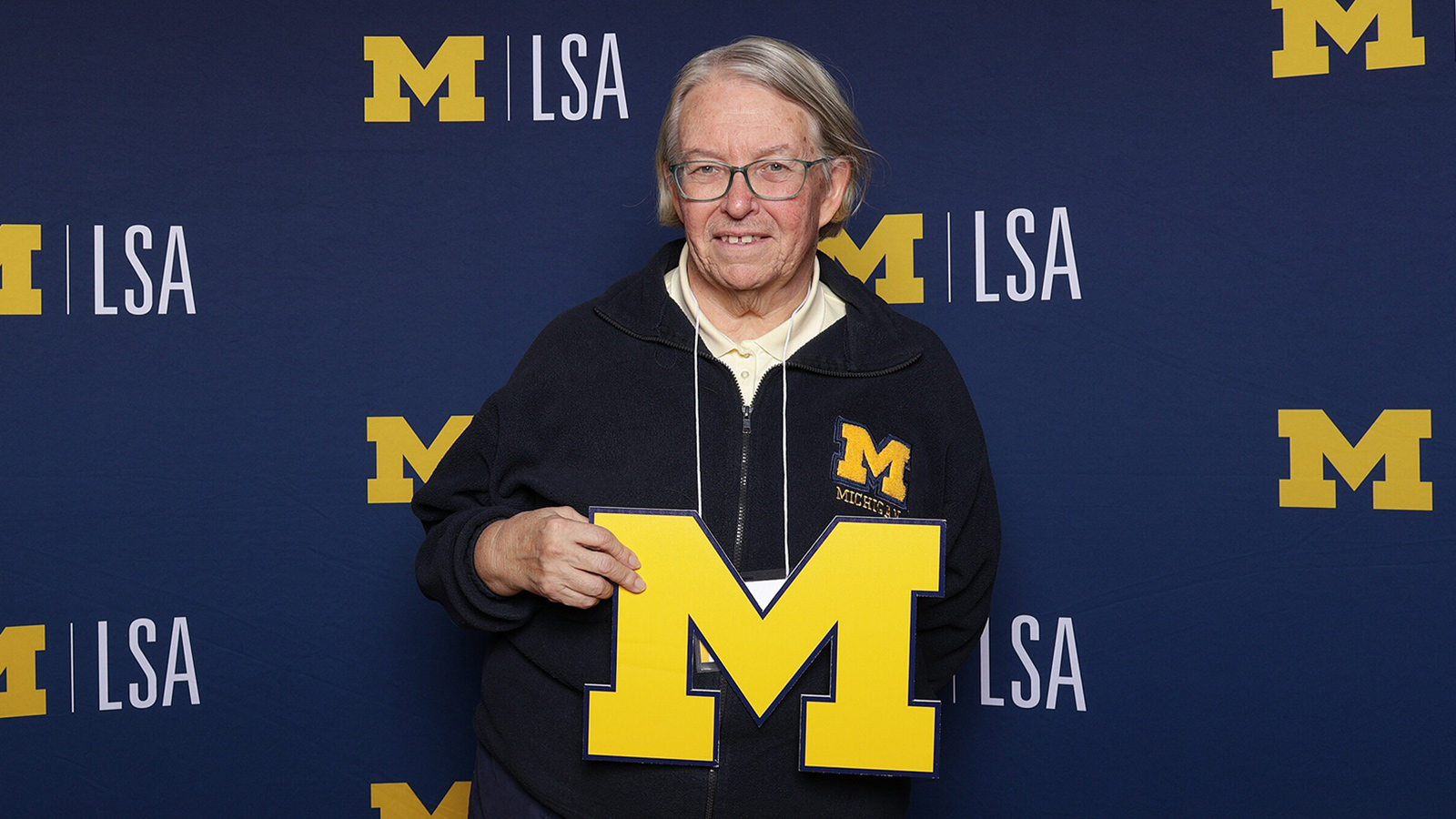 A woman holding a yellow block M, standing in front of a backdrop that has yellow block M’s followed by the letters LSA displayed in white.