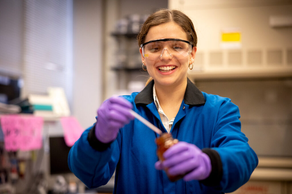 A smiling female student in a lab coat, rubber gloves, and protective goggles handling lab equipment.