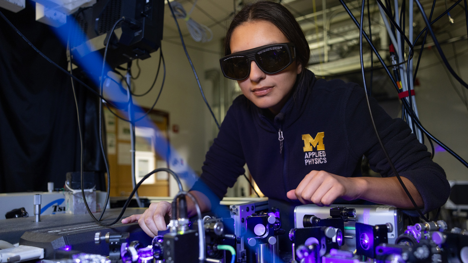 A person wearing dark glasses working with equipment surrounded by wires and purple light