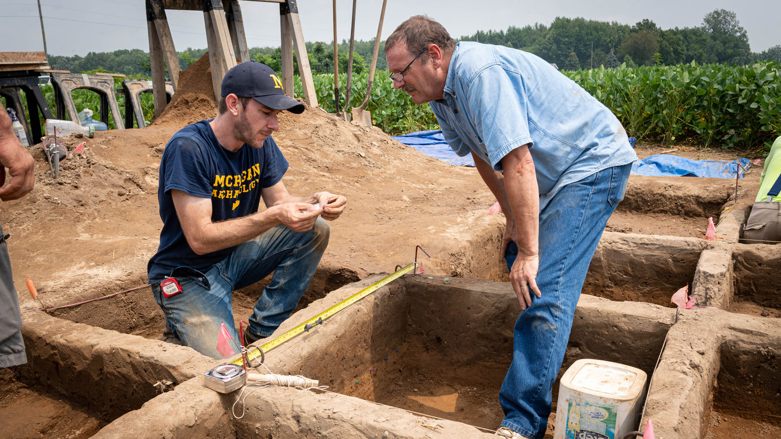 Two people are working at an archaeological dig site. The site is marked with pink flags, tools, and a white container, with fields and trees in the background.