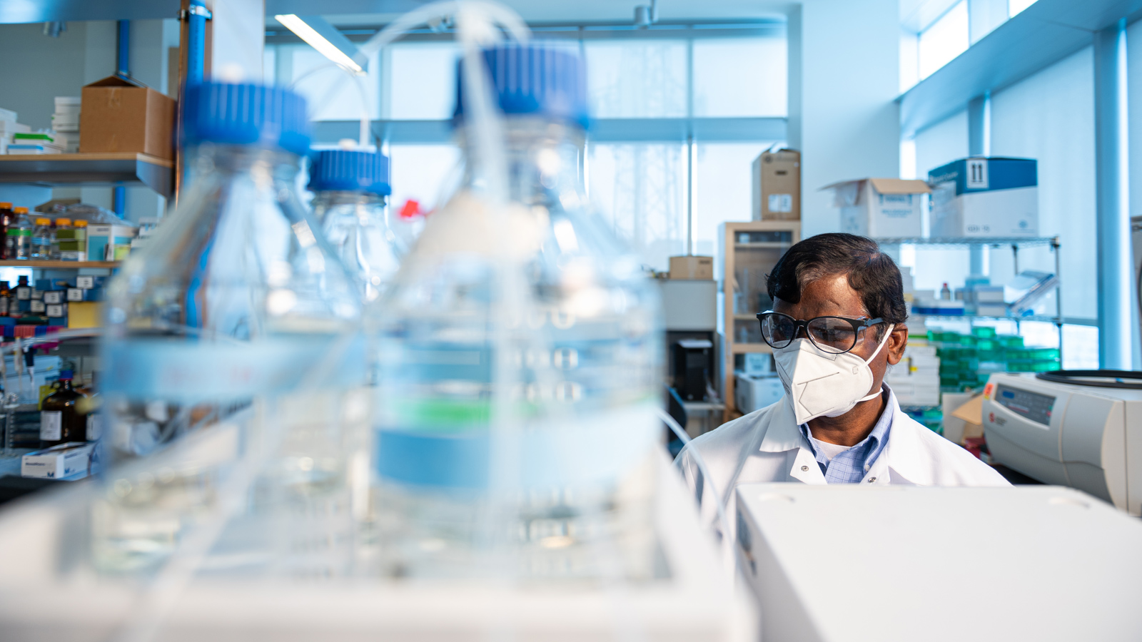 A researcher wearing a lab coat in a laboratory filled with scientific equipment, including bottles with blue caps and shelves stocked with supplies.