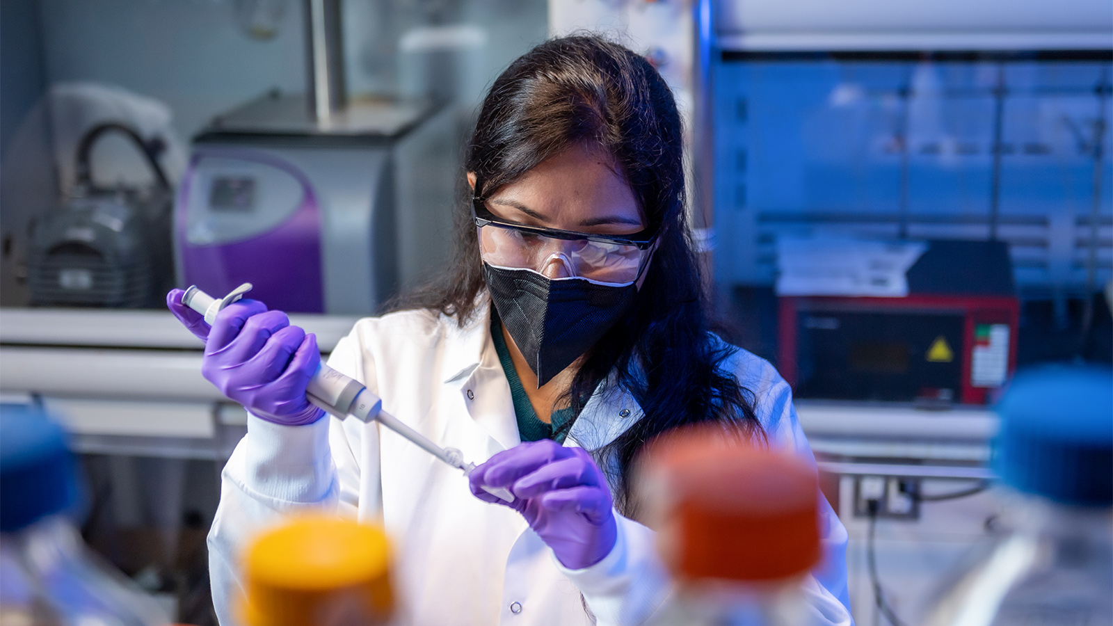 A person wearing a lab coat and purple gloves uses a pipette in a laboratory, surrounded by various bottles and scientific equipment.