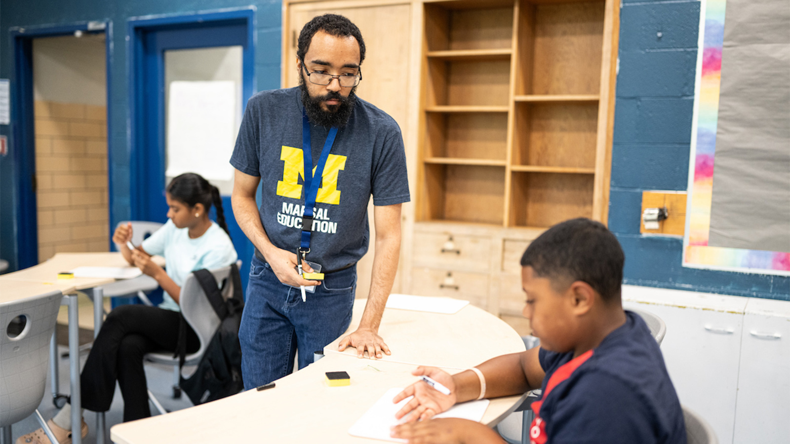 A teaching intern wearing a University of Michigan Marsal Education shirt assists a student with an activity at a classroom table.