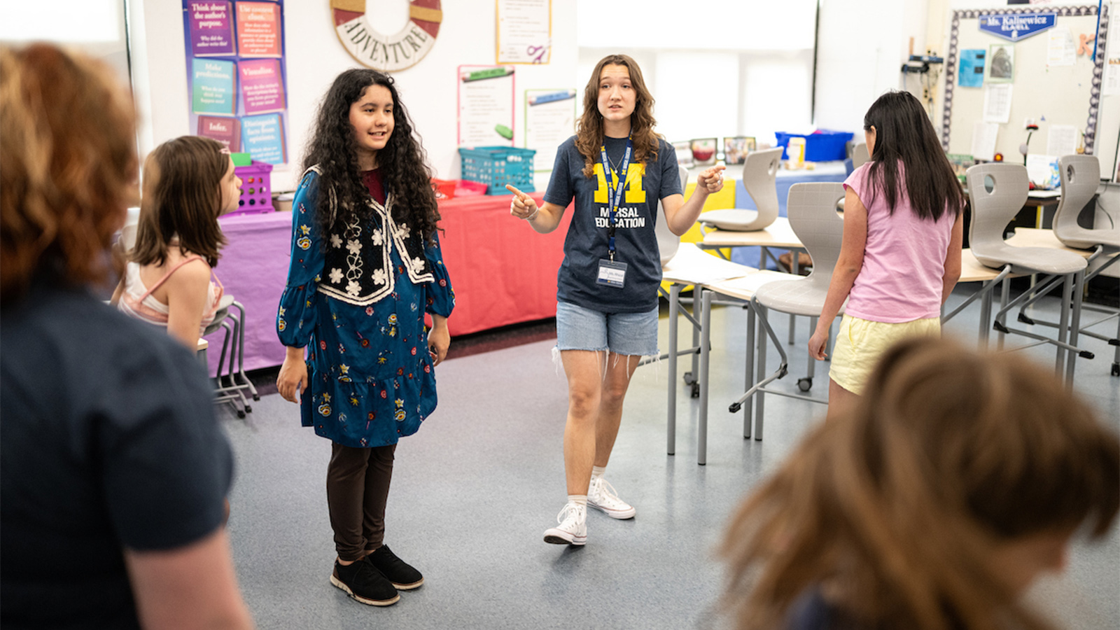 A teaching intern wearing a University of Michigan Marsal Education shirt, leads a group activity with children in a classroom.