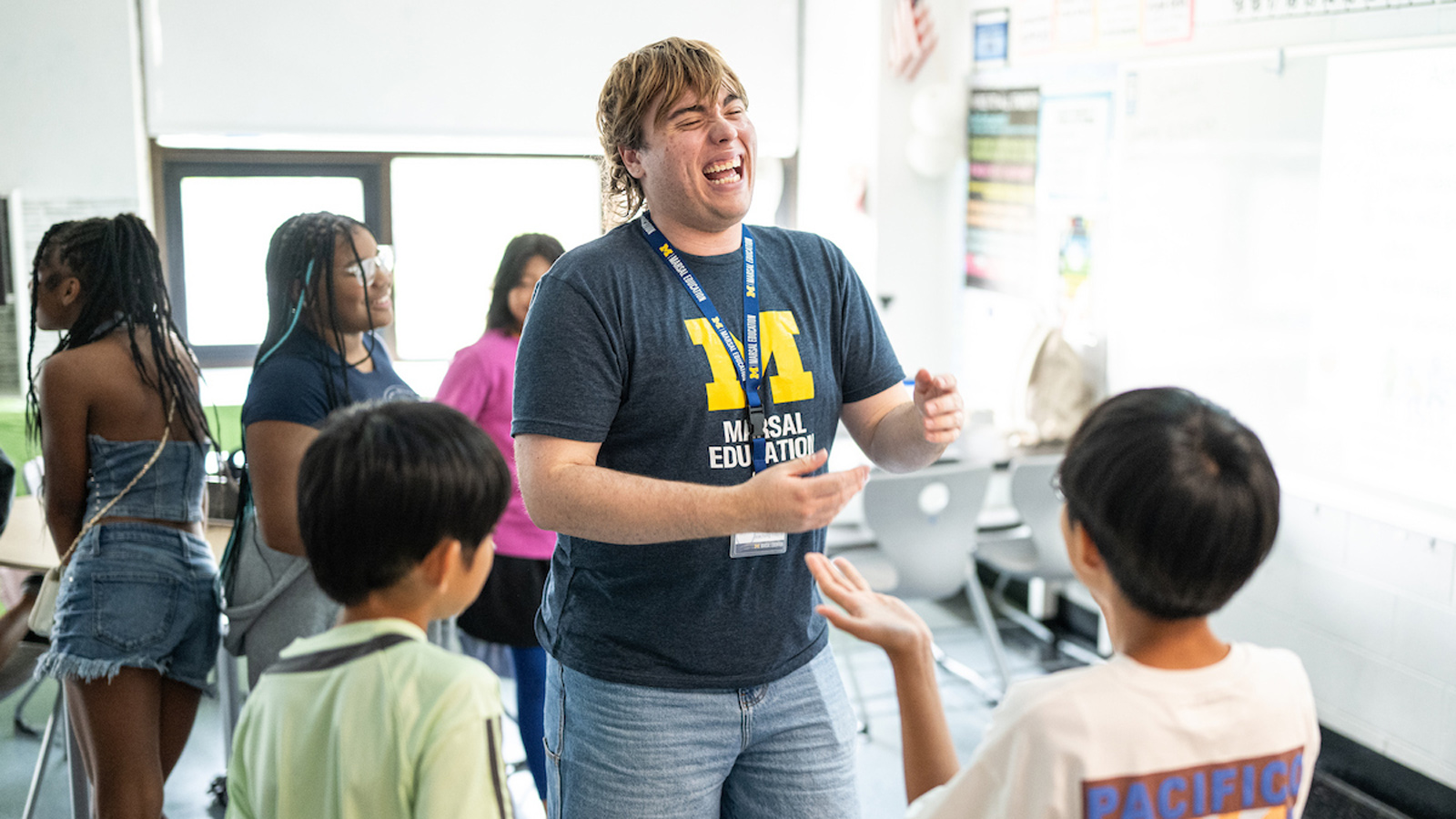 A smiling teaching intern wearing a University of Michigan Marsal Education t-shirt engages with two students in a bright, active classroom.