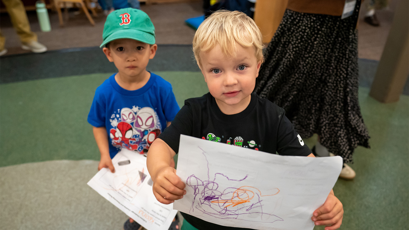A young child proudly holds up a colorful scribble drawing while another child in a green Boston Red Sox baseball cap stands nearby with a paper.