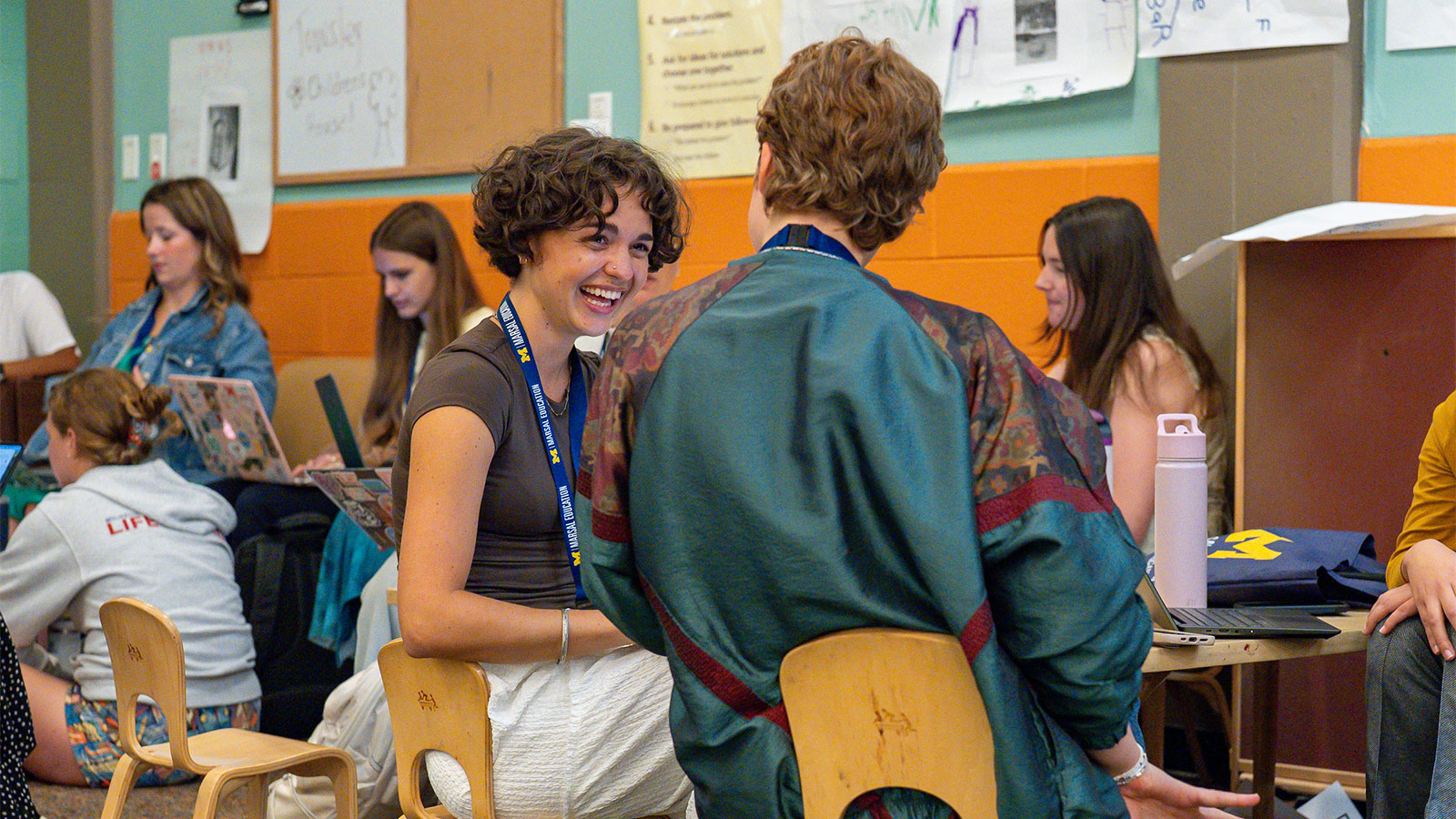 A person smiles broadly while talking with another person in a lively classroom gathering.