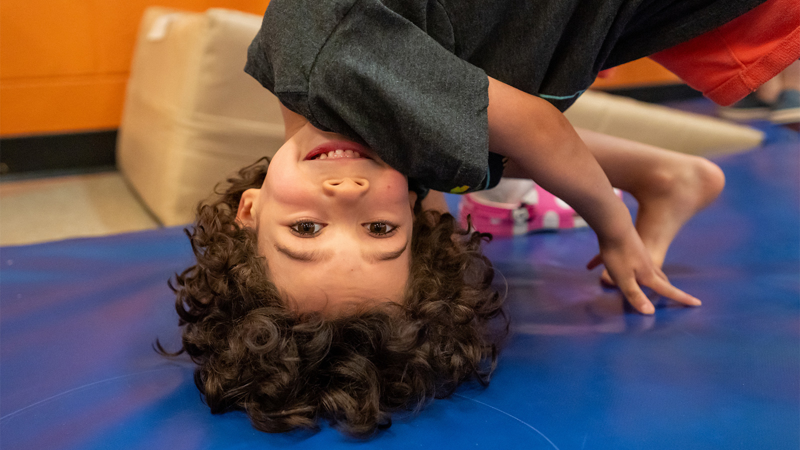A child smiles while hanging upside down over a blue padded mat during a movement activity.