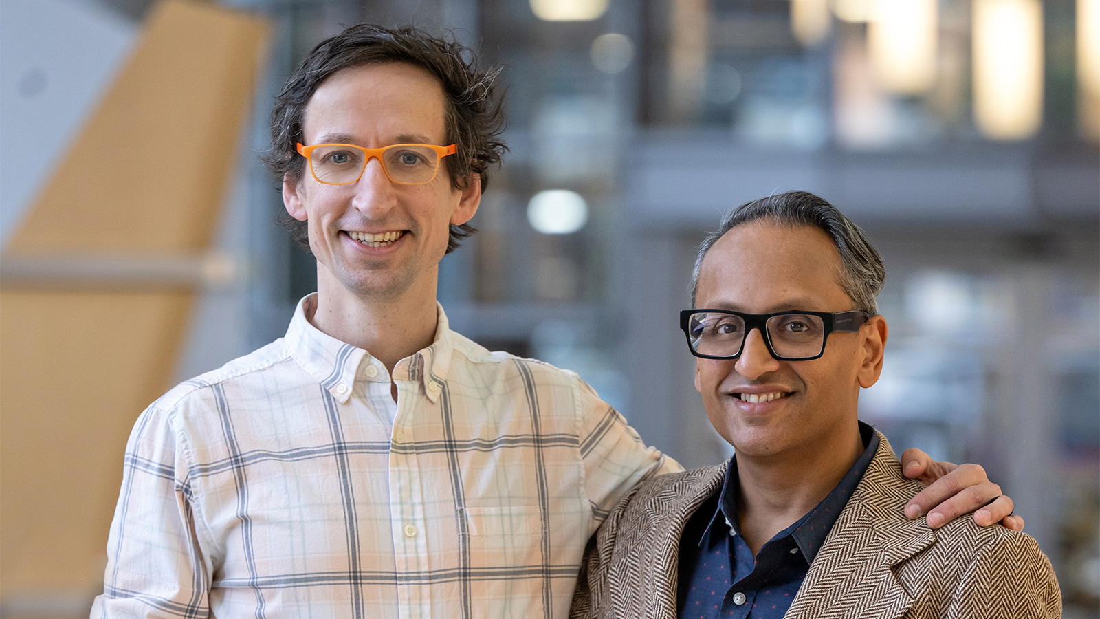 Two men wearing glasses and business casual attire standing next to each other with an arm around one another. 