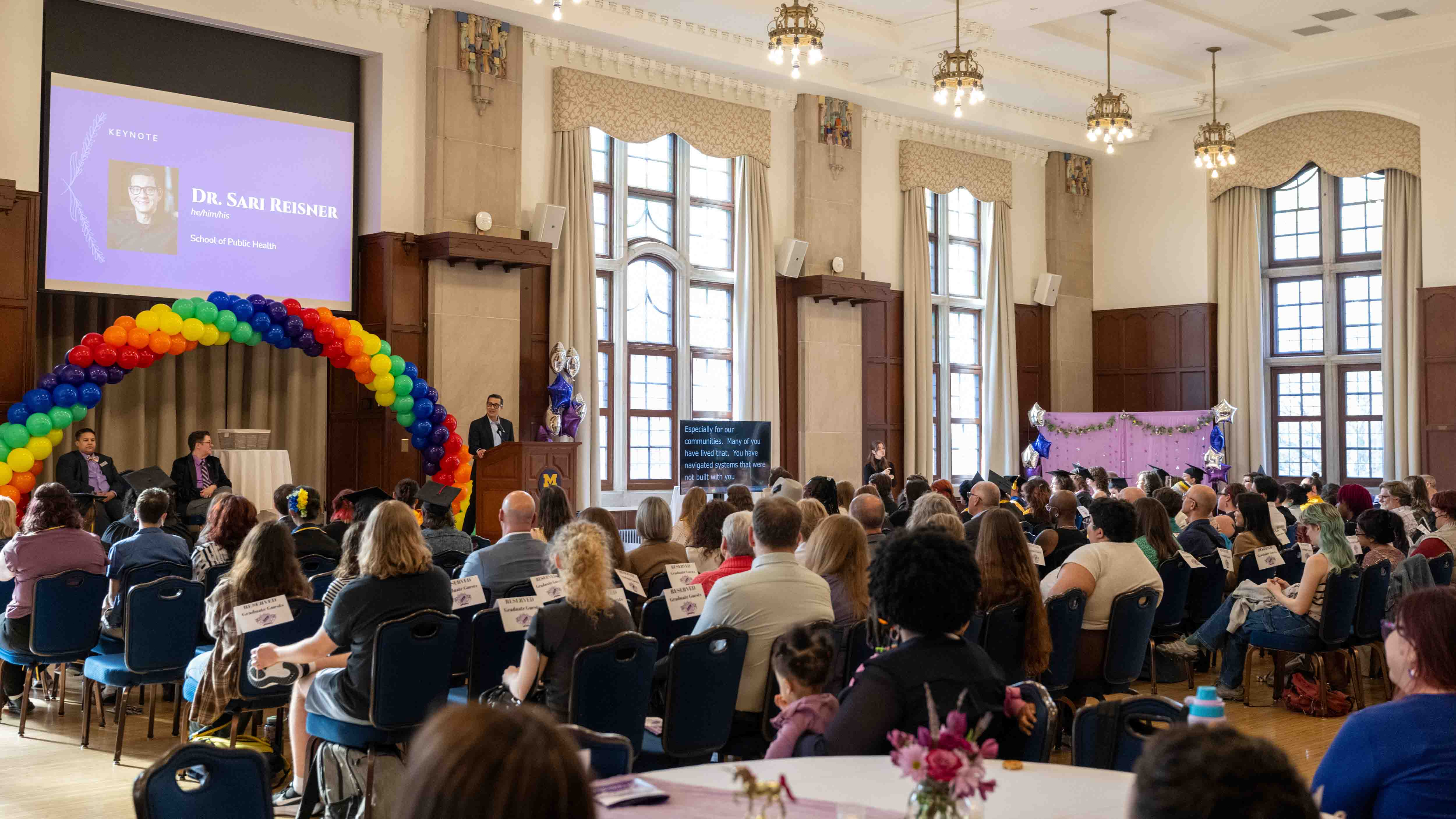 A view of the audience from the back of the Rogel Ballroom at Lavender Graduation. The room is decorated with a rainbow balloon arch by the speaker’s podium and a lavender photo backdrop in the corner.
