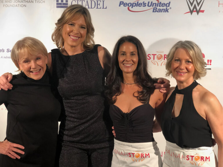 Joyce, Storm, Wolfson, and Kreamer stand in front of a background with the names of different event sponsors written on it.