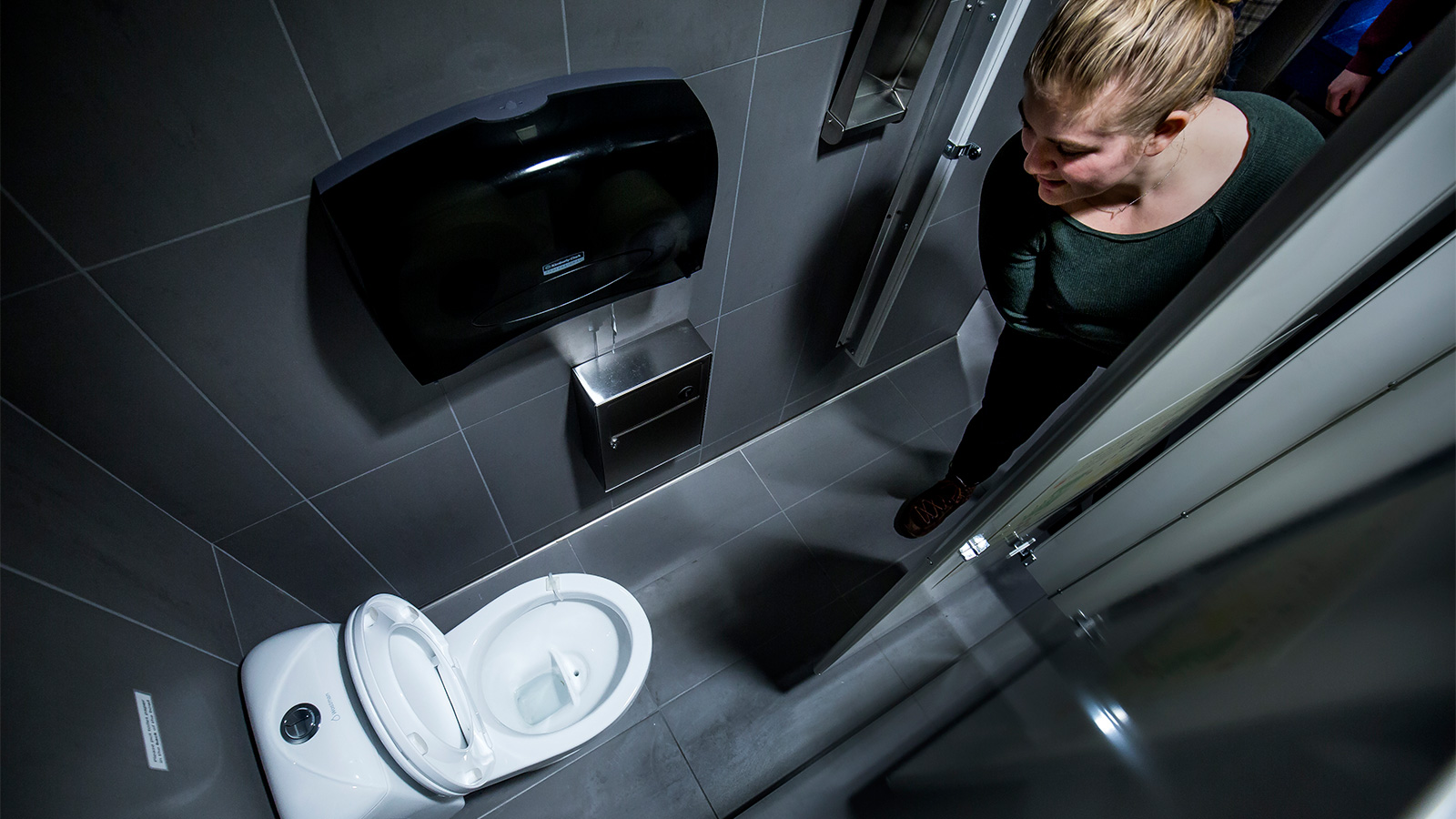 A person looks into a compact restroom stall with an open toilet, gray tiled walls and floor, and wall-mounted dispensers.